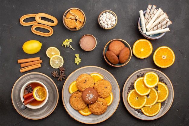 Top view yummy sugar cookies with cup of tea and oranges on dark background sugar tea biscuit cookie sweet fruit