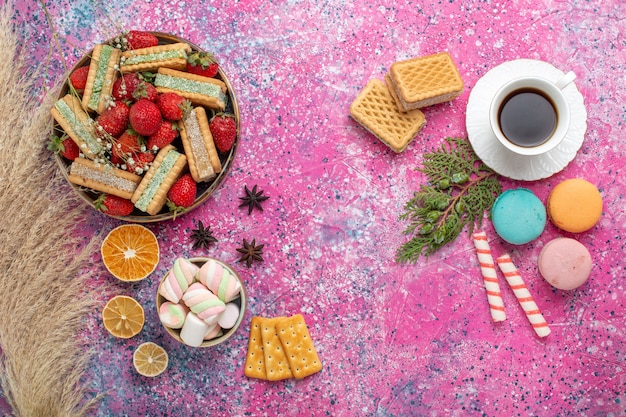 Top view of yummy waffle cookies with french macarons and fresh red strawberries on pink surface