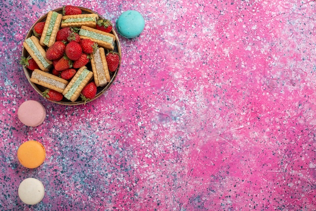 Вид сверху вкусного вафельного печенья со свежей красной клубникой и французскими макаронами на розовой поверхности
