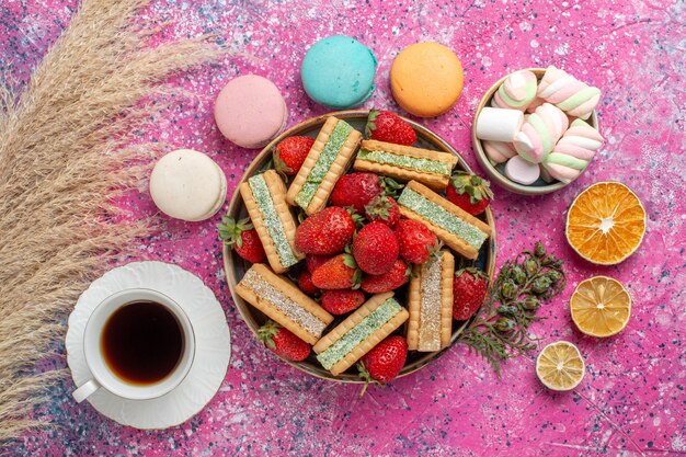 Top view of yummy waffle cookies with fresh red strawberries macarons and tea on pink surface