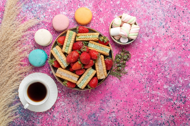 Вид сверху вкусного вафельного печенья со свежими красными макаронами из клубники и чаем на розовой поверхности