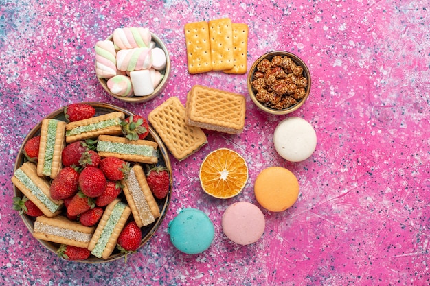 Vista dall'alto di deliziosi biscotti waffle con marshmallow macarons e fragole rosse fresche sulla superficie rosa