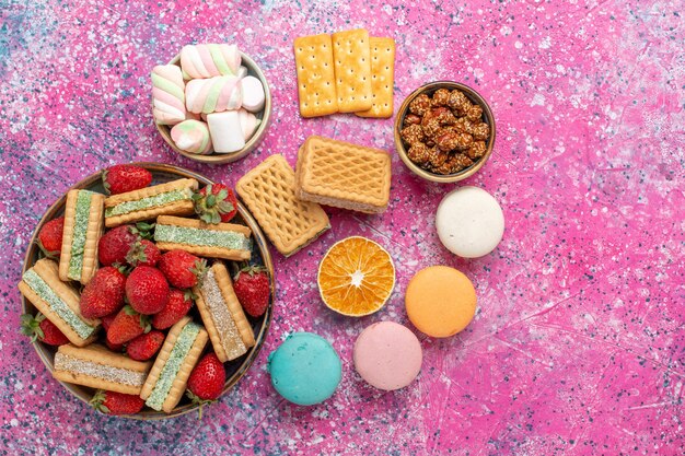 Top view of yummy waffle cookies with marshmallows macarons and fresh red strawberries on pink surface