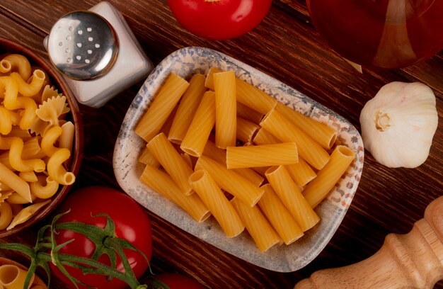 Top view of ziti pasta with different types in bowl and salt tomato garlic on wooden surface