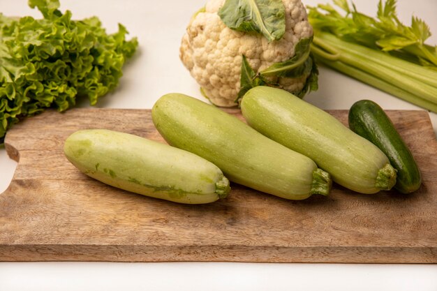 Top view of zucchinis on a wooden kitchen board with cucumbers lettuce celery and cauliflower isolated on a white surface