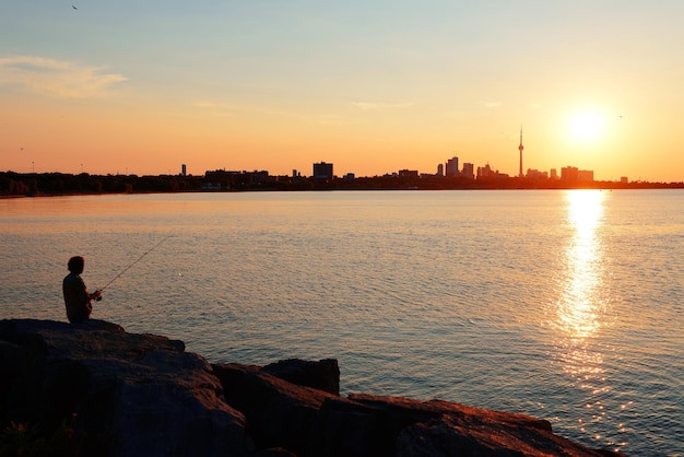 Toronto sunrise silhouette over lake with man fishing.