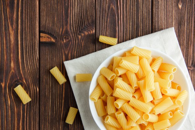 Tortiglioni pasta in a bowl on wooden and kitchen towel background, flat lay.