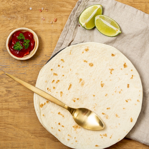 Tortilla with spoon, tomato sauce and sliced lime on table