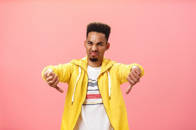 Totally dislike it is awful. Disgusted and displeased african american male with beard and afro haircut frowning pursing lips, grimacing from dissatisfaction showing thumbs down gesture over pink wall