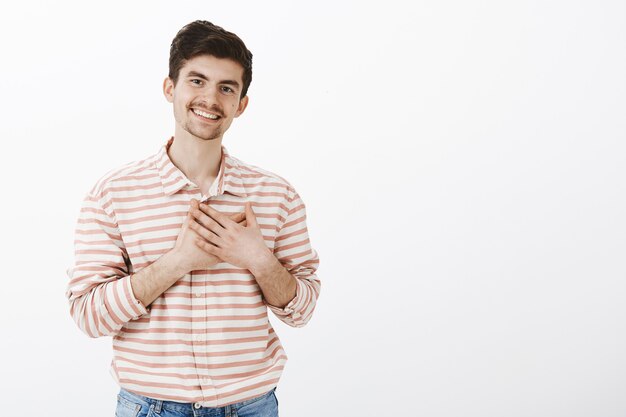 Touched to bottom of heart. Indoor shot of pleased caring handsome boyfriend with moustache and beard, holding hands on heart and smiling lovely, feeling warmth after hearing loving words