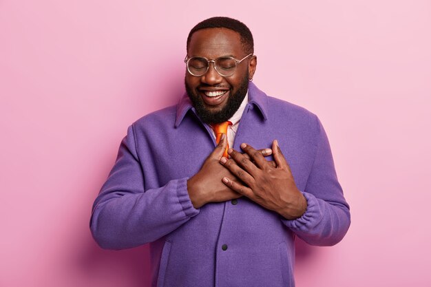 Прикосновенный счастливый бородатый чернокожий мужчина слышит трогательные слова, выражает доброту, носит прозрачные очки, носит оптические очки, фиолетовый пиджак