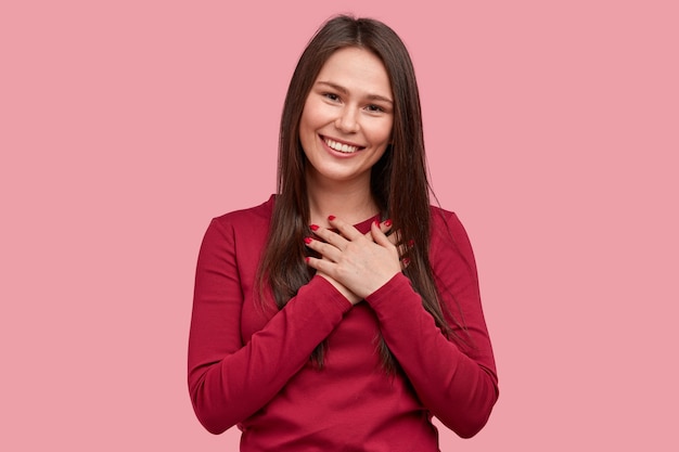 Touched positive woman with pleased expression keeps hands on chest, feels gratitude, impressed by good words of gratitude, isolated over pink background. People