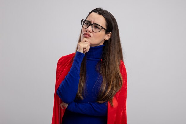 toughtful young superhero girl looking at side wearing glasses putting hand on chin isolated on white