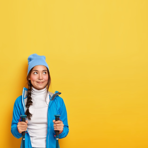 Концепция туризма и кемпинга. Мечтательная брюнетка-женщина использует треккинговые палки, смотрит вверх, наслаждается активным отдыхом, задумчиво смотрит вверх, носит синюю куртку и шляпу, копирует пространство на желтой стене.