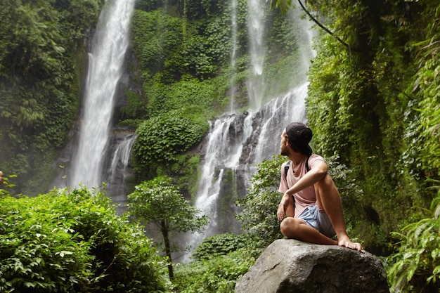 Туризм, путешествия и приключения. Стильный молодой хипстер сидит на камне босиком и поворачивает голову, чтобы увидеть удивительный водопад