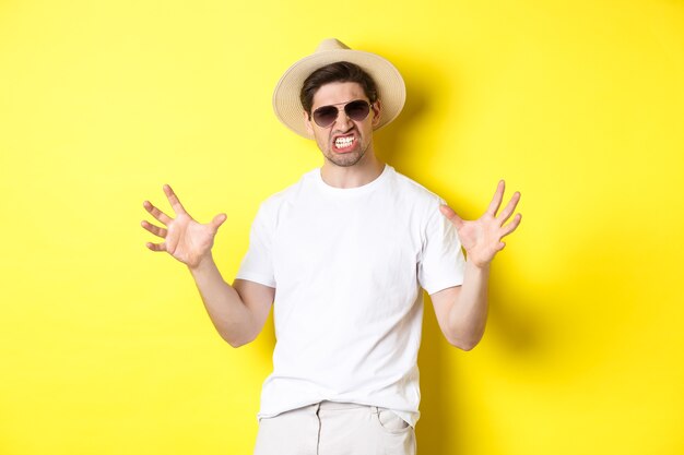 Концепция туризма, путешествий и праздников. Нахальный молодой человек в отпуске, показывая что-то большое и стиснув зубы, стоит в солнечных очках и летней шляпе.