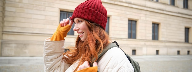 Free photo tourism and travelling young redhead woman smiling tourist walking with backpack around city centre