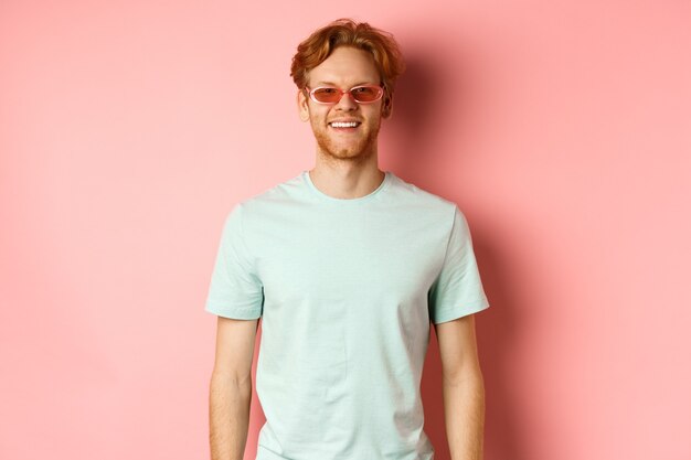 관광 및 휴가 개념입니다. 선글라스와 티셔츠를 입은 쾌활한 빨간 머리 수염 남자, 웃고 분홍색 배경 위에 서 있는 카메라를 보고 행복해 보인다