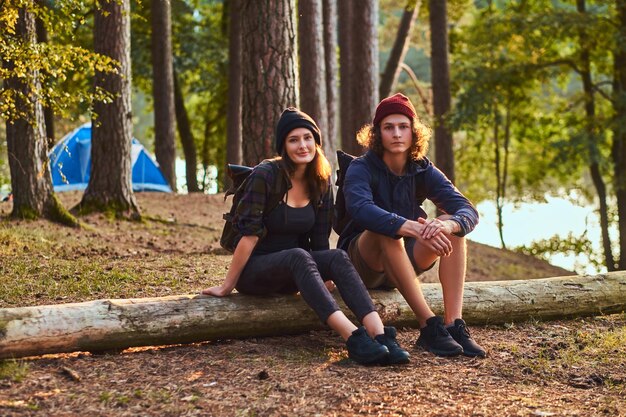 Tourist girl holds hand on chin while sitting on a tree trunk and holds a smartphone in the forest.