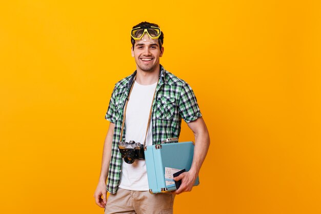 Tourist guy wearing plaid shirt and white T-shirt looking at camera. Portrait of man with diving mask on his head, holding retro camera and hand suitcase.