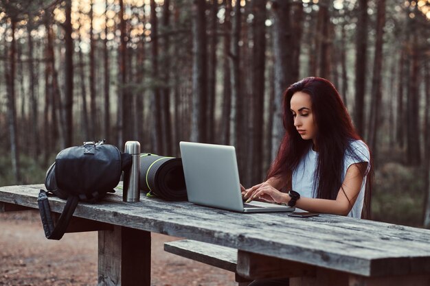 Tourist hipster girl in white shirt working on a laptop while sitting on a wooden bench in the beautiful autumn forest.