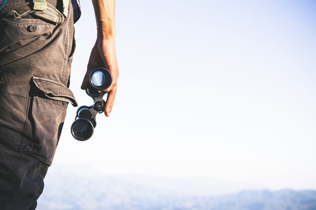 Tourist is holding through binoculars on sunny cloudy sky from mountain top.