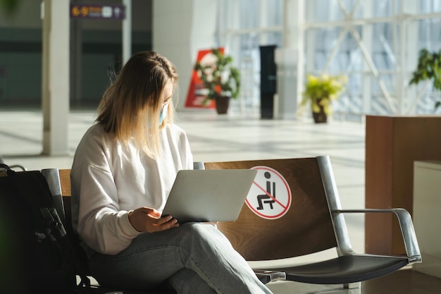 사회적 거리 표지판이 있는 공항 로비 의자에 앉아 노트북을 사용하는 관광객