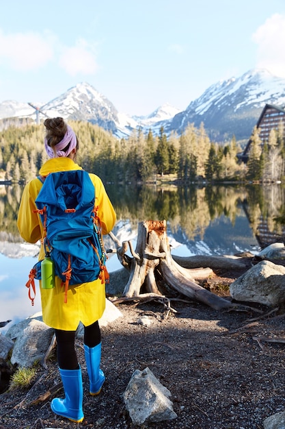 Tourist with backpack stands near mountain river, enjoys wild nature with beautiful view, wears yellow long anorak