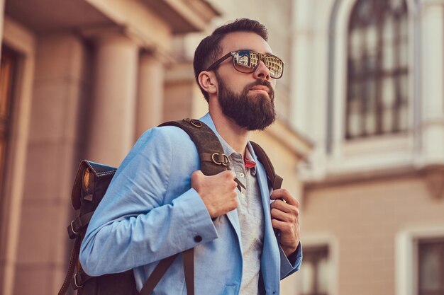 A tourist with a full beard and haircut, wearing casual clothes and sunglasses, holds a backpack, standing on an antique street, during the excursion in Europe.