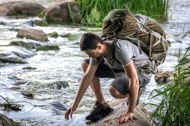 Un turista con un grande zaino da trekking si sta rinfrescando vicino a un fiume di montagna nella calura estiva.
