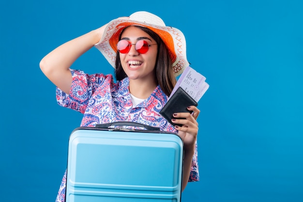 彼女の帽子に触れる幸せと肯定的な顔に笑顔で旅行スーツケースとチケットとパスポートを保持している赤いサングラスをかけた帽子を持つ観光女性