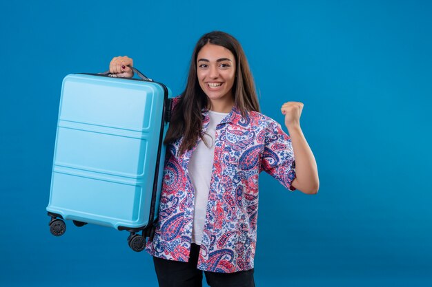 旅行スーツケース探しの観光客女性は彼女の成功と彼女の目的と分離された青の上に立って目標を達成するために幸せな喜びで彼女の拳を握りしめている勝利を喜んで終了しました
