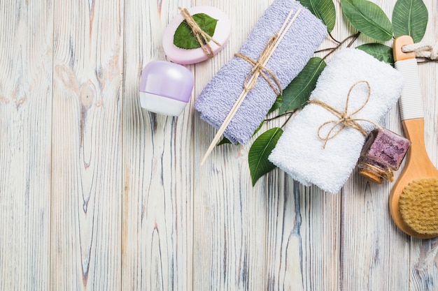 Towels; scrub bottle; moisturizing cream; leaves; brush and soap on wooden plank