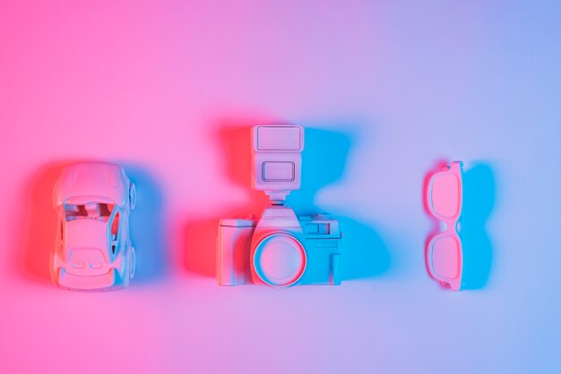 Toy car; retro camera and spectacle arranged in a row on pink backdrop with blue light effect