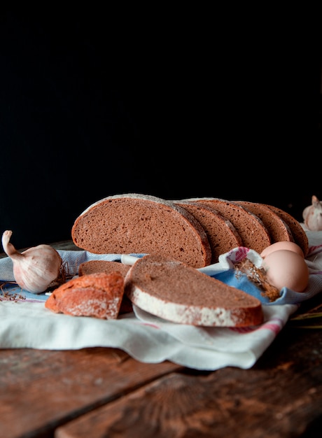 Traditional black bread side view thin slices with white flour on it, on a rustic towel  with garlic and eggs, on a wooden kitchen table.