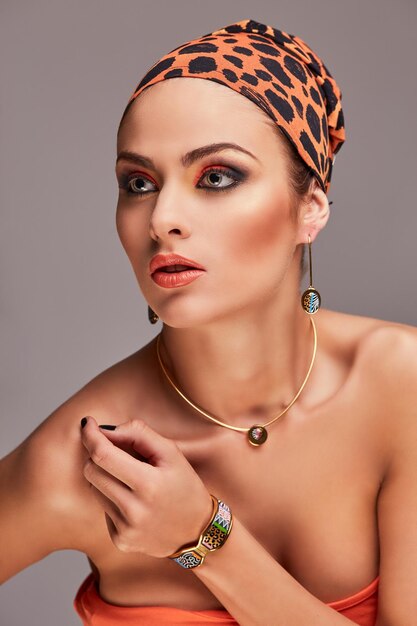 Traditional fashion beauty. Sensual caucasian woman wearing colorful shawl on head and accessories, posing with hand on shoulder. Isolated on gray background.