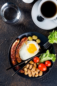 Традиционный полный английский завтрак с яичницей, сосисками, фасолью, помидорами на тарелке, круассаном и кофе на темном фоне. вид сверху