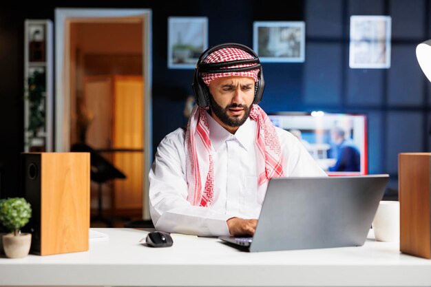 Традиционный ближневосточный предприниматель усердно работает в современном офисе, используя беспроводные технологии для общения и исследований. Молодой араб пользуется беспроводными наушниками и ноутбуком.
