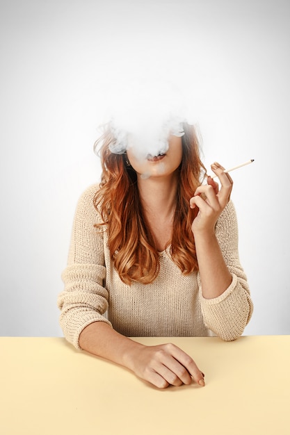 Tranquil woman sitting and smoking resting at the table. Cloud of smoke covering her face. Copy space