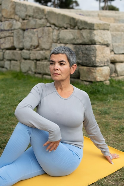 Free photo tranquil woman with grey hair doing yoga in park. female model in sportive clothes exercising on mat on cloudy day. sport, hobby, health concept