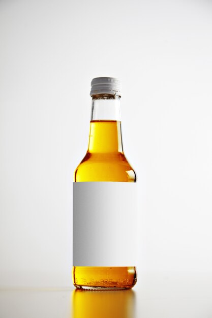 Прозрачная стеклянная запечатанная бутылка, изолированная на простом фоне с белой пустой этикеткой и вкусным напитком внутри