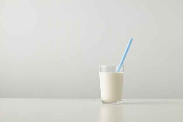 Transparent glass with fresh organic milk and blue drinking straw inside isolated on side of white table