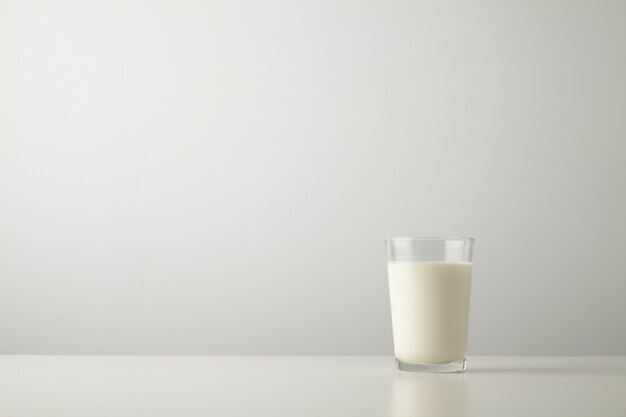 Transparent glass with fresh organic milk isolated on side of white table. Space for your text above