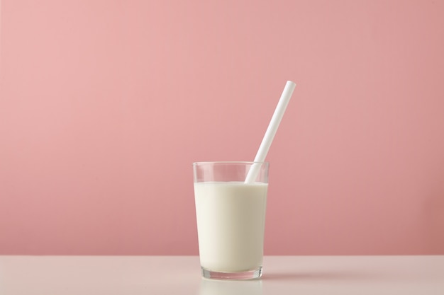 Transparent glass with fresh organic milk and white drinking straw inside isolated on pastel pink background on wooden table