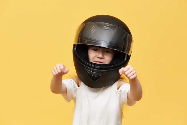 Транспорт, экстрим, автоспорт и концепция деятельности. Портрет опасной маленькой девочки-наездницы в черном защитном мотоциклетном шлеме, держащей руки перед собой, как будто за рулем мотоцикла