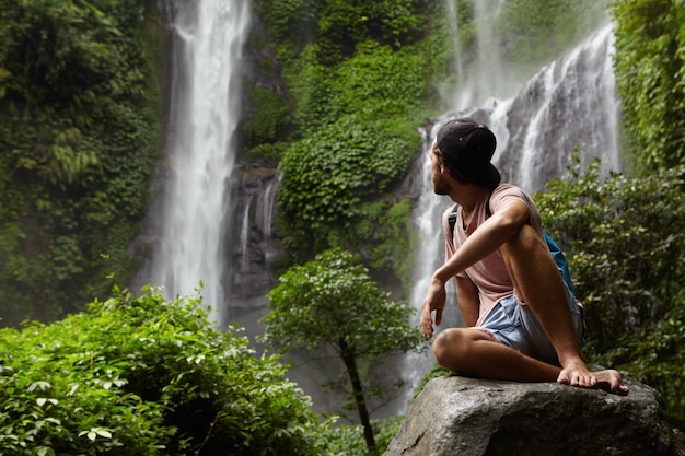 旅行と冒険。スナップバックと石の上に座って、美しい緑の熱帯雨林の滝を振り返ってバックパックを着ておしゃれな若い男。ジャングルの岩の上に残りの部分を持つ裸足の観光客