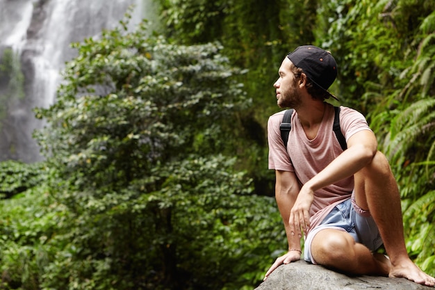 Путешествие и приключения. Красивый молодой босоногий турист-мужчина с рюкзаком расслабляется в одиночестве на большом камне и оглядывается назад