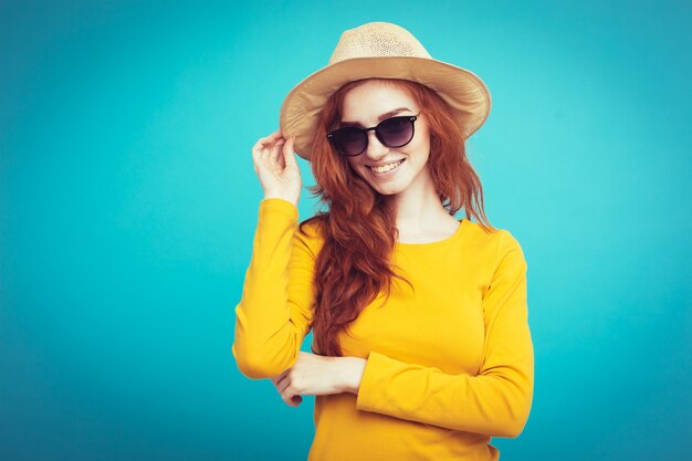 旅行の概念 - クローズアップ肖像画若い美しい魅力的な赤毛の女の子と流行の帽子とサングラス笑顔。青いパステルの背景。スペースをコピーします。