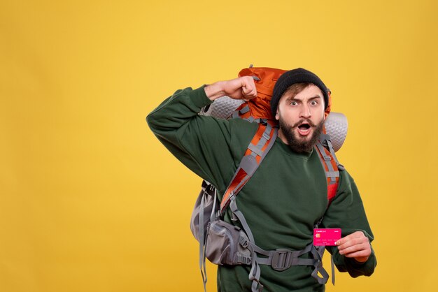 Travel concept with ambitious proud young guy with packpack and holding bank card on yellow 