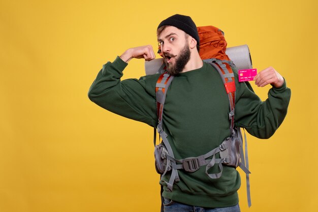 Travel concept with ambitious young guy with packpack and holding bank card showing muscular on yellow